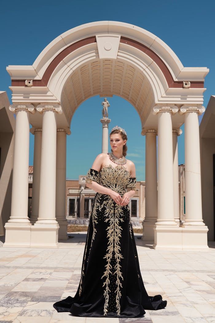 An elegant woman poses in a luxurious black gown with gold embellishments, framed by classical architecture.