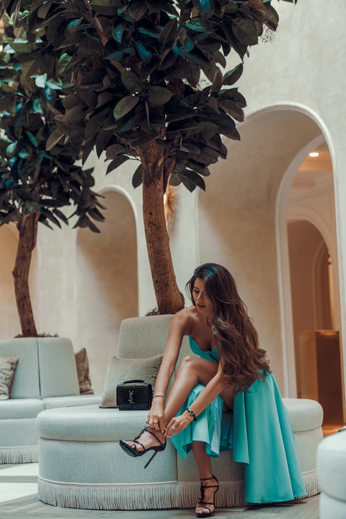 Stylish woman in turquoise dress adjusting heels in a luxurious indoor setting.