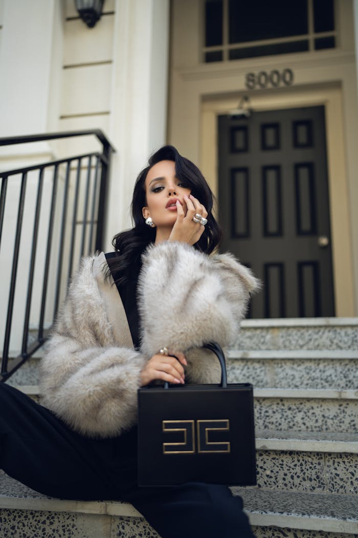 Fashionable woman in fur coat poses with luxury handbag on city steps.